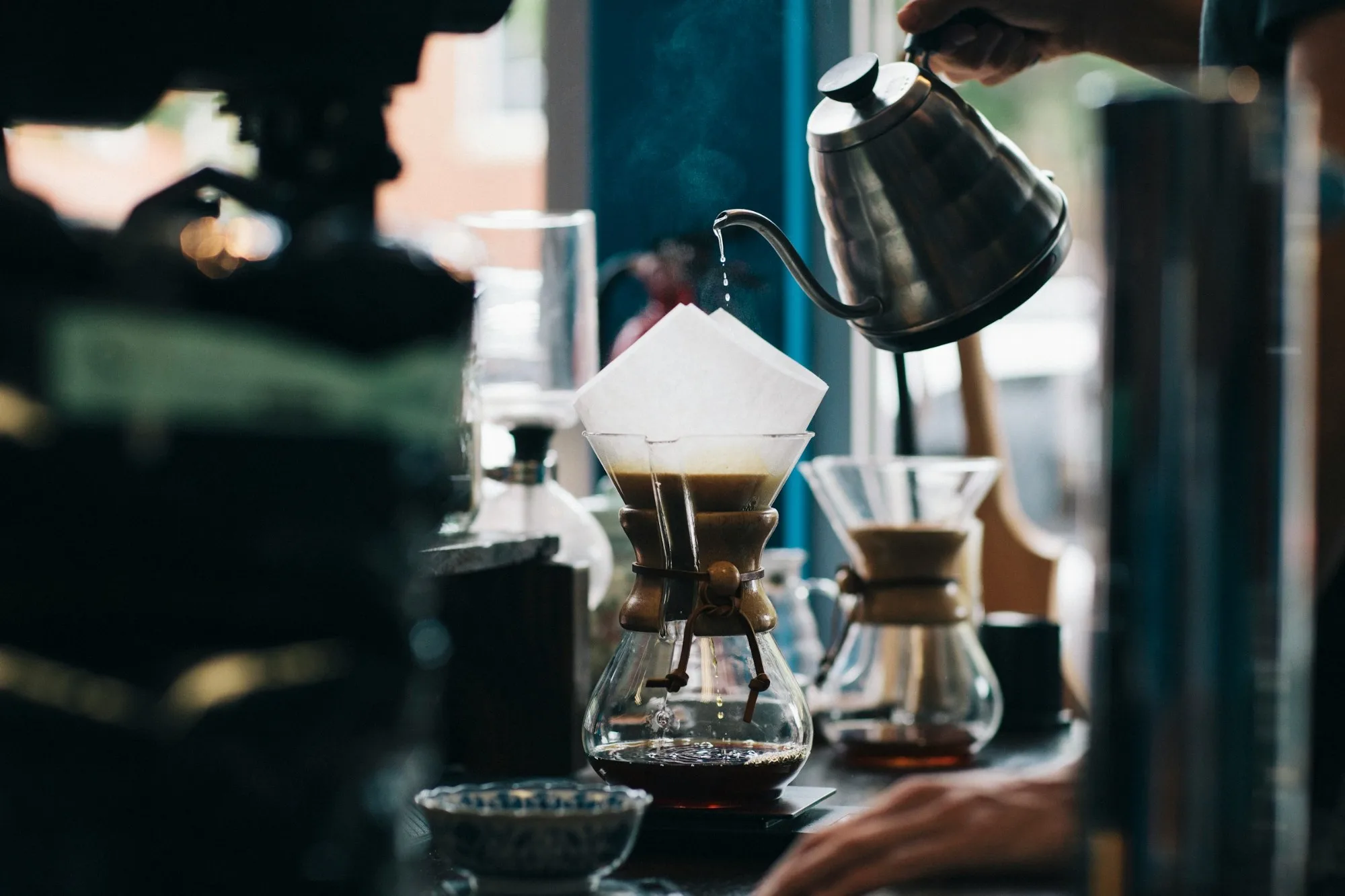 A barista pouring hot water from a gooseneck kettle into a Chemex pour-over coffee brewer on a cafe counter, with a second brewer beside it