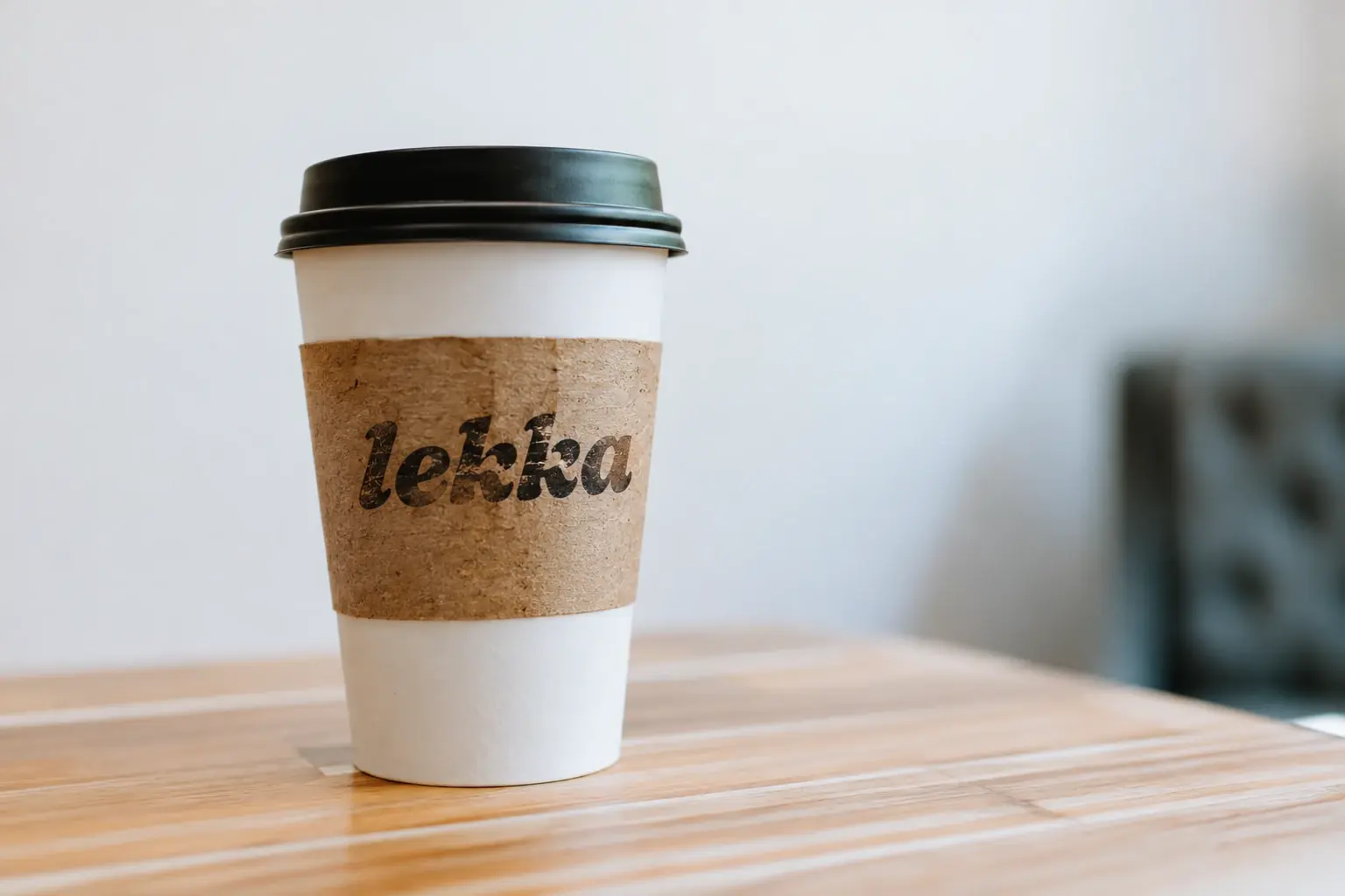 A takeaway coffee cup in a brown sleeve stamped lekka, standing on a wooden cafe table.